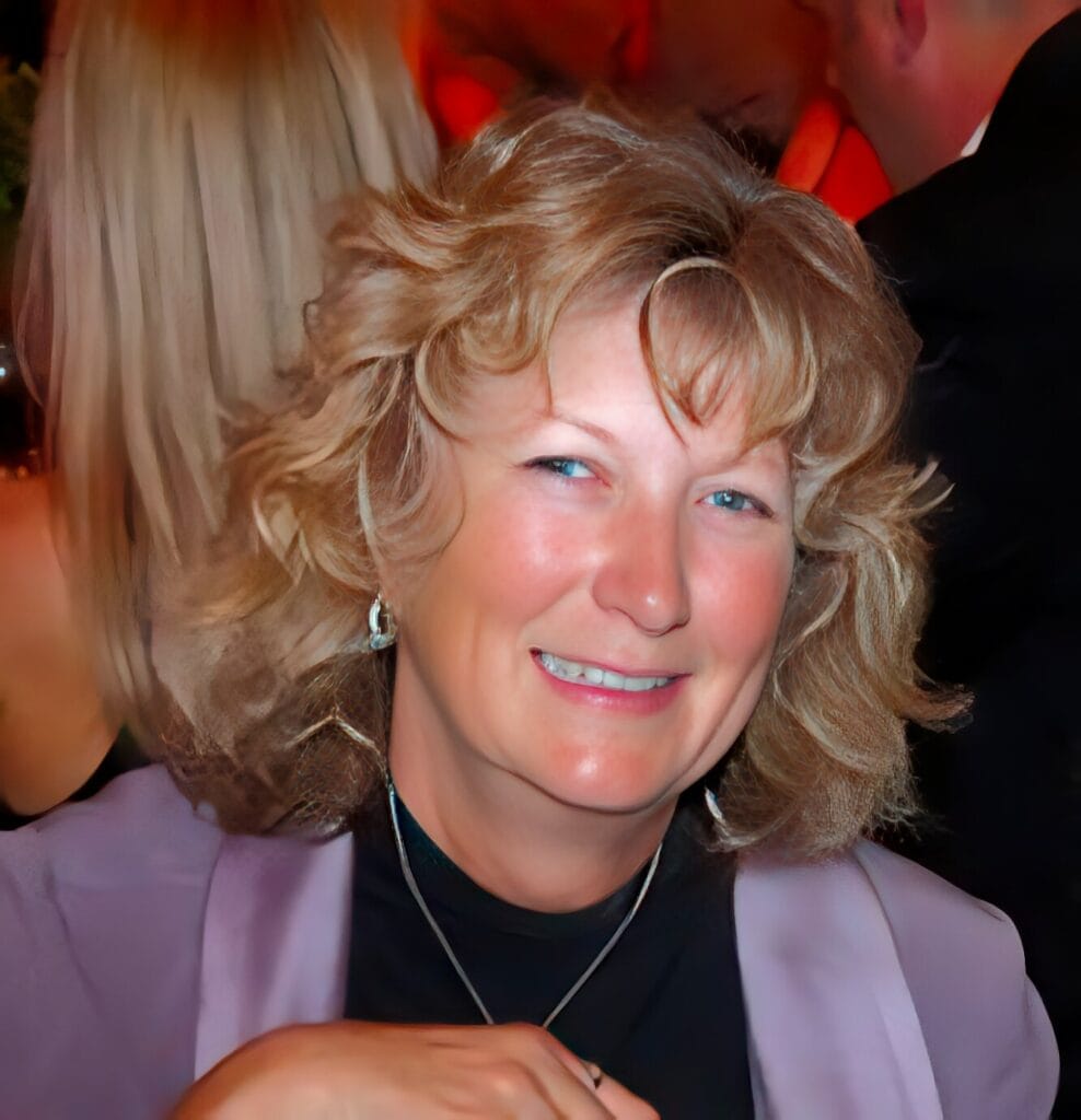 JillAlchorn A woman with wavy blonde hair, wearing a lavender blazer and black top, smiles while seated indoors at a social event.