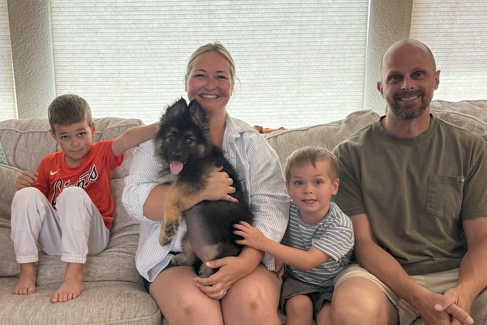 Lucy Family2 A family of four sits on a couch with a German Shepherd puppy; two boys are seated on either side of a smiling woman holding the dog, and a man sits at the end.